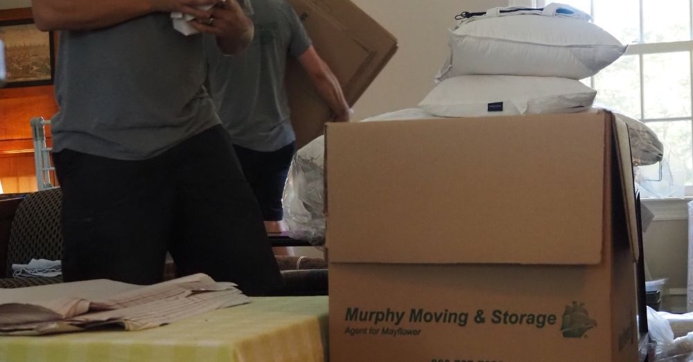 Two movers carry boxes and other items in a room. A large "Murphy Moving & Storage" box is in the foreground.