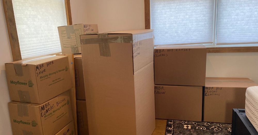 Stack of labeled cardboard moving boxes in a room with white blinds.