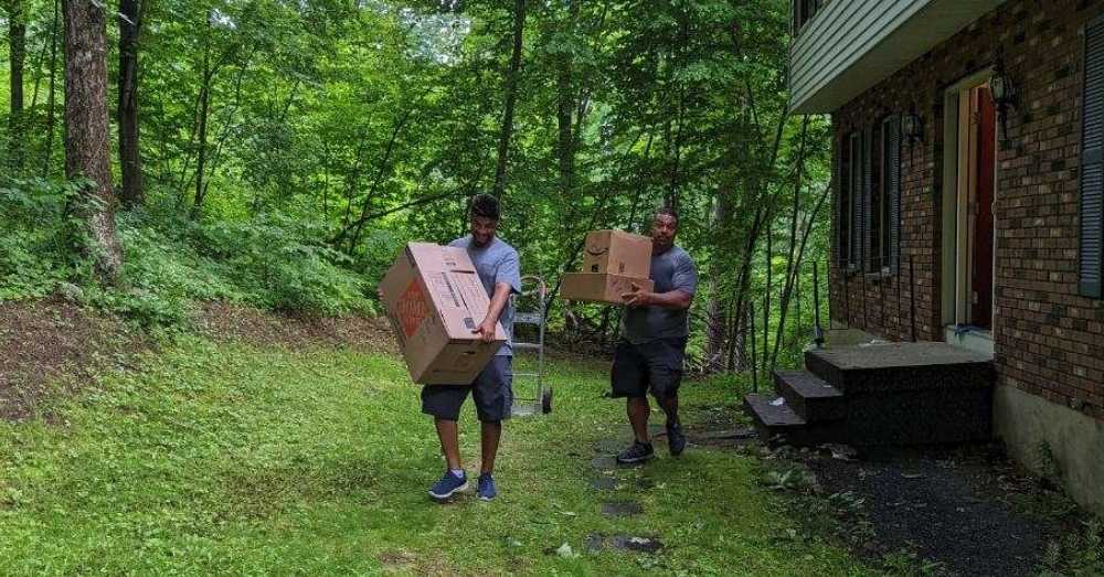 Two people moving boxes—one large, one small stack—out of a house surrounded by a green forest.