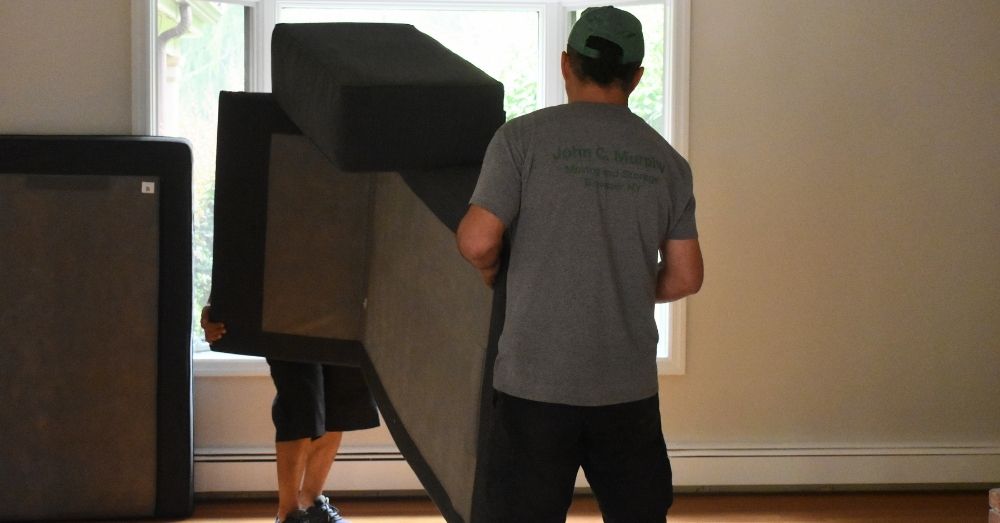 Two movers carrying a large, dark piece of furniture, likely a box spring or mattress, through an empty room.