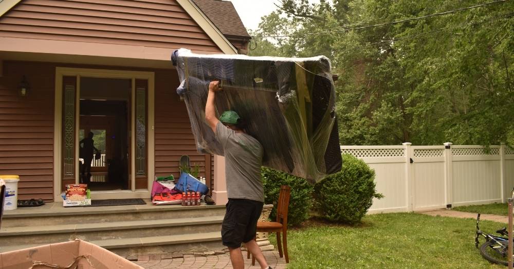 A mover carries a large wrapped piece of furniture out of a house, prepping to move in Hartford CT