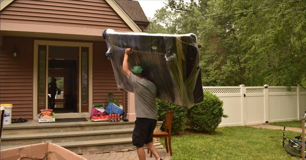 A mover carries a large wrapped sofa out of a house.