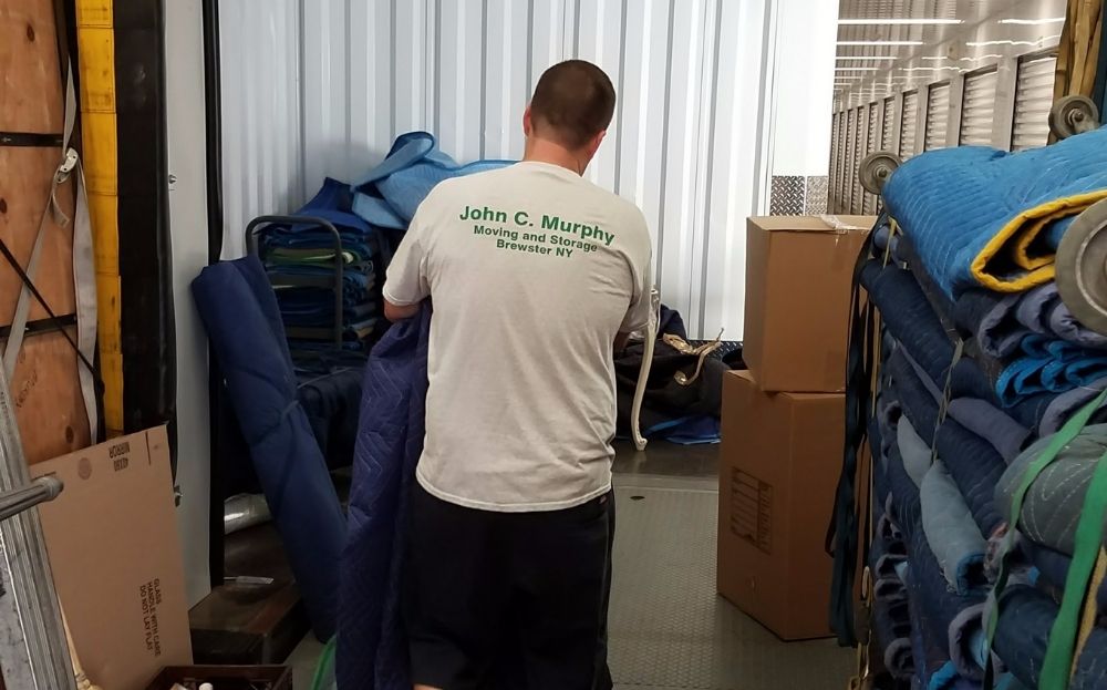 A man wearing a John C. Murphy Moving Storage shirt is organizing moving blankets and boxes inside a portable storage unit.