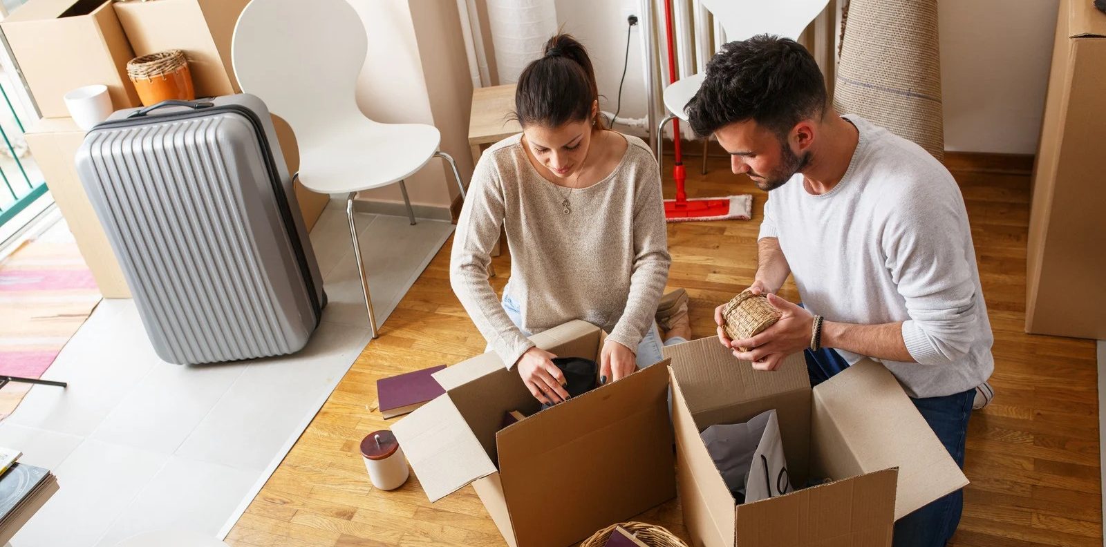 A man and woman kneeling on a wooden floor in a room filled with boxes, selectively packing small household items into cardboard containers. A large silver suitcase and various furniture pieces are visible in the background, illustrating the final packing and preparation phase two days before a move.