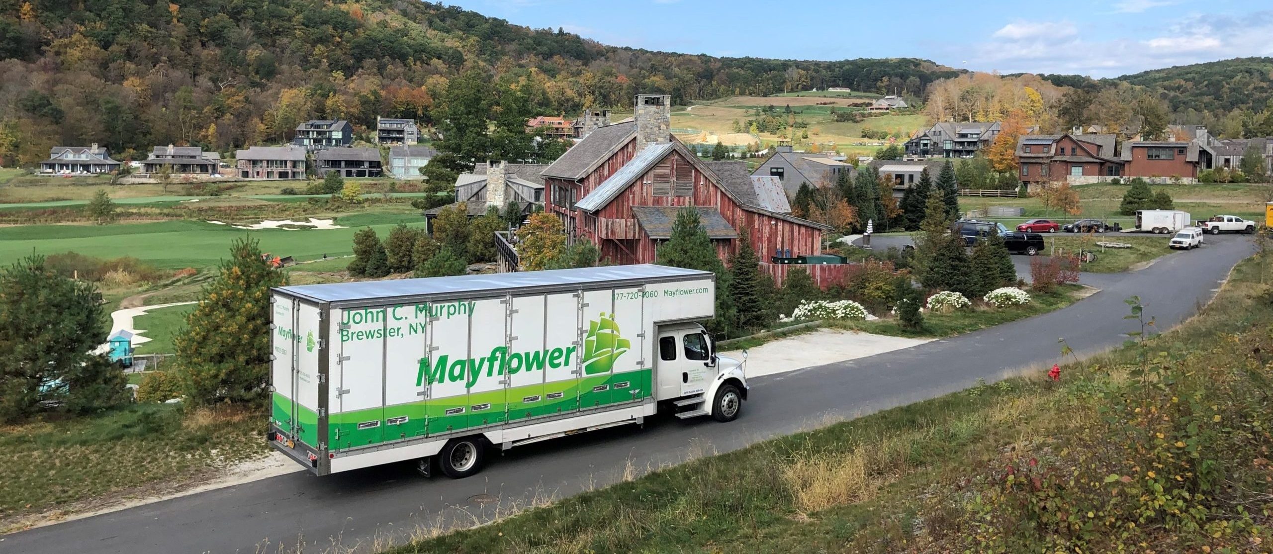 A wide, elevated rear-view shot of a white and green Murphy Moving & Storage truck traveling down a winding asphalt road in Amenia, NY. The background features rolling hills, lush green trees, and several large gray residential homes under a bright, partly cloudy sky, illustrating the coordination required for long-distance moves.