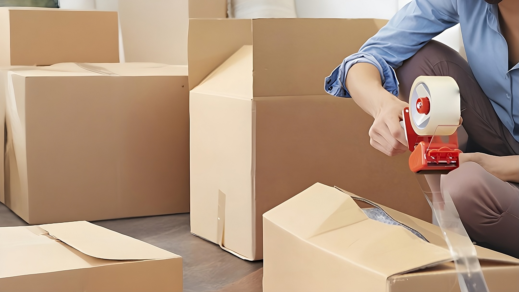 A close-up view of a person’s hand using a red handheld tape dispenser to seal the top of a brown cardboard moving box. Other boxes are visible in the blurred background, highlighting the physical labor involved in packing for a move.