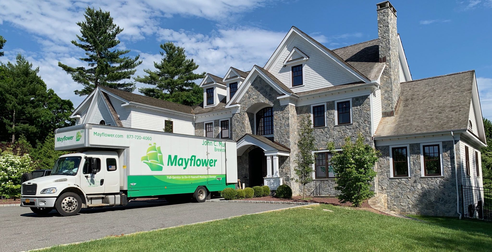 A wide shot of a white and green Mayflower moving truck parked on a gray asphalt driveway in front of a luxury two-story estate. The home features a combination of stone and white siding, surrounded by a large green lawn and pine trees, representing a strategically timed weekday move.