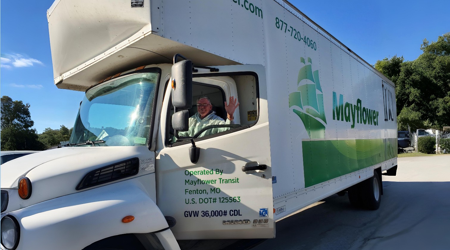A close-up of "Danny," a Murphy professional driver, smiling and waving from the cab of a white and green Mayflower moving truck. The truck features contact information and DOT numbers, representing a reputable and verified moving service.