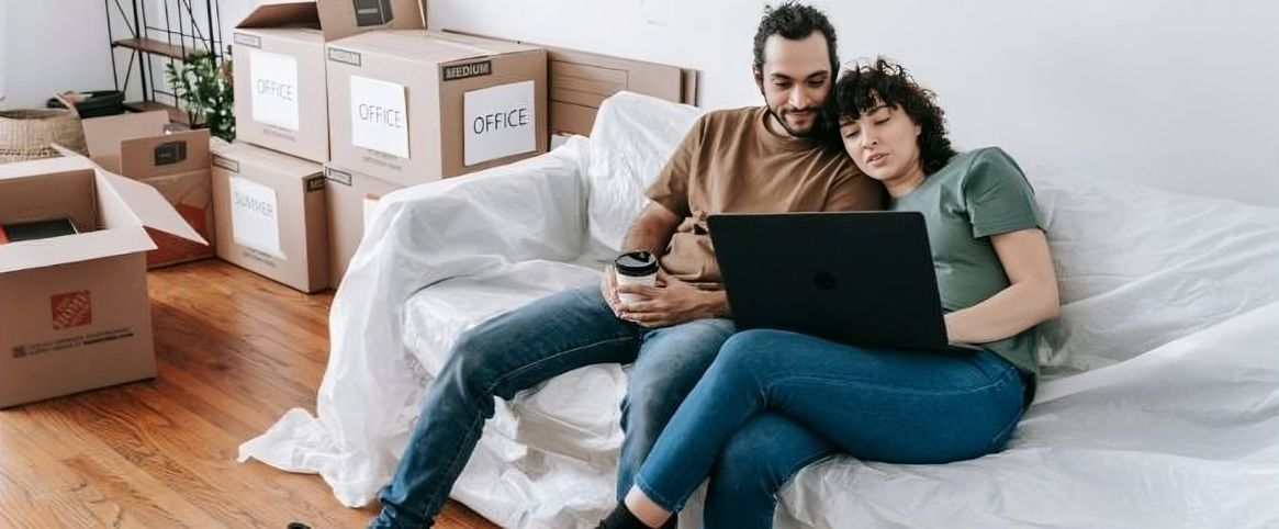 A couple sits on a couch with a laptop, surrounded by moving boxes.