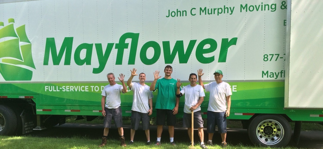 Five moving crew members wave and smile while standing in front of a Mayflower moving truck