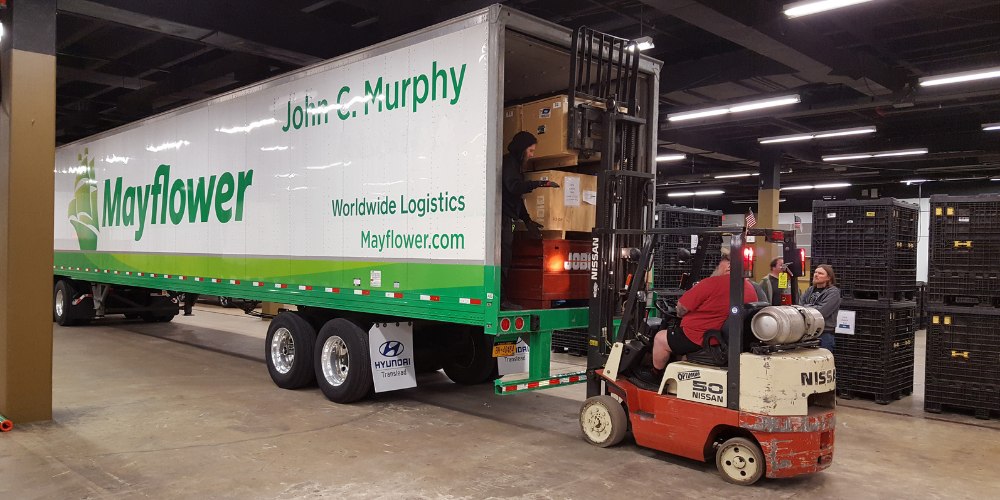 A forklift operator lifts large wooden cargo crates into the back of a Mayflower moving semi-truck trailer