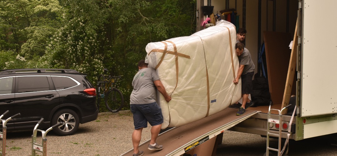 Three movers pushing a wrapped mattress up a ramp