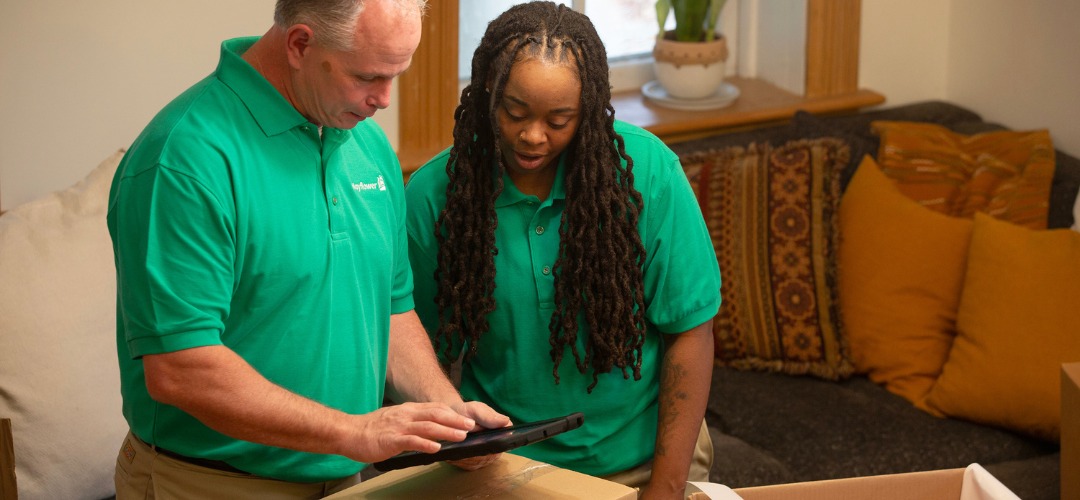 two people wearing green polo shirts look at a tablet over moving boxes